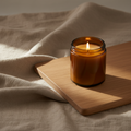 Lit candle in an amber glass jar on linen beside a pale wooden board